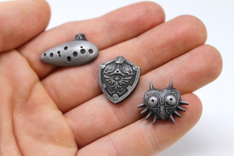 May include: Three silver-coloured metal pins featuring iconic symbols from the Legend of Zelda video game series. The pins depict the Ocarina of Time, the Hylian Shield, and Majora's Mask.