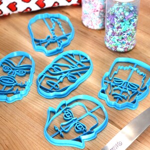May include: Six light blue plastic cookie cutters in various skull designs are arranged on a wooden surface. Two clear plastic jars filled with colorful sprinkles are in the background. A blue spatula is on the right.