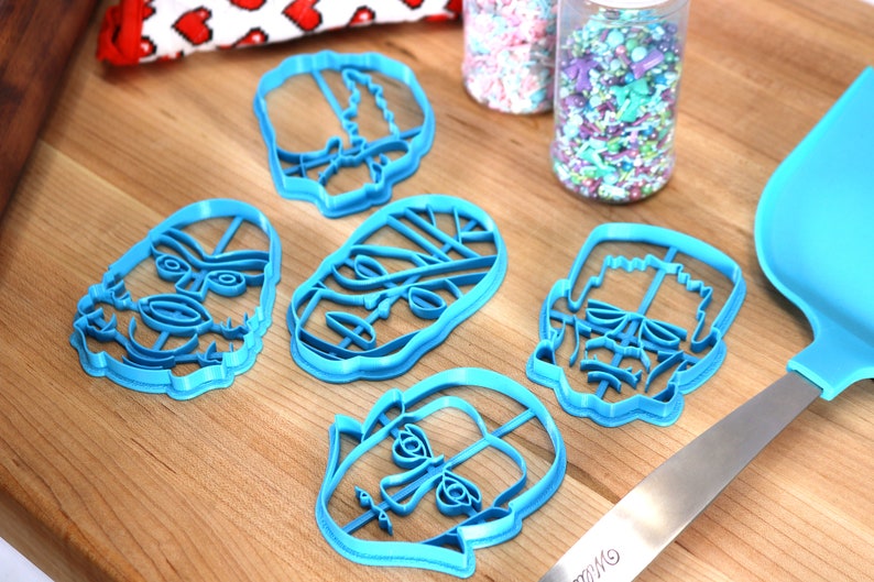 May include: Six light blue plastic cookie cutters in various skull designs are arranged on a wooden surface. Two clear plastic jars filled with colorful sprinkles are in the background. A blue spatula is on the right.