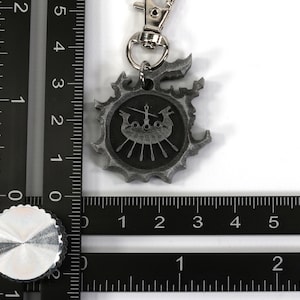 Op de afbeelding: Een zilverkleurige metalen sleutelhanger met een gestileerd schip met riemen en een zwaard, omringd door een cirkelvormig ontwerp met een gekartelde rand.