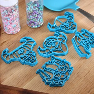 May include: A set of six blue plastic cookie cutters shaped like a skull with horns. The cutters are on a wooden surface with two jars of sprinkles in the background.