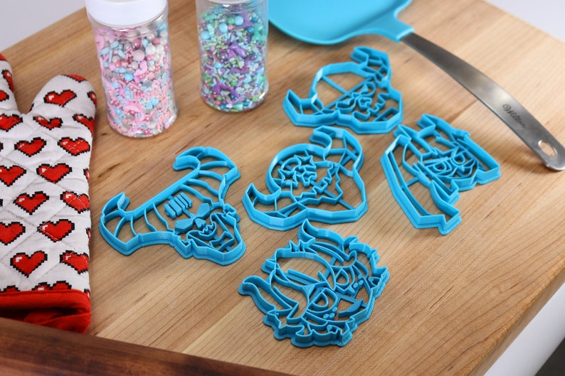 May include: A set of six blue plastic cookie cutters shaped like a skull with horns. The cutters are on a wooden surface with two jars of sprinkles in the background.