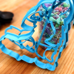 May include: A blue plastic cookie cutter shaped like a mermaid, with a jar of colourful sprinkles in the background.
