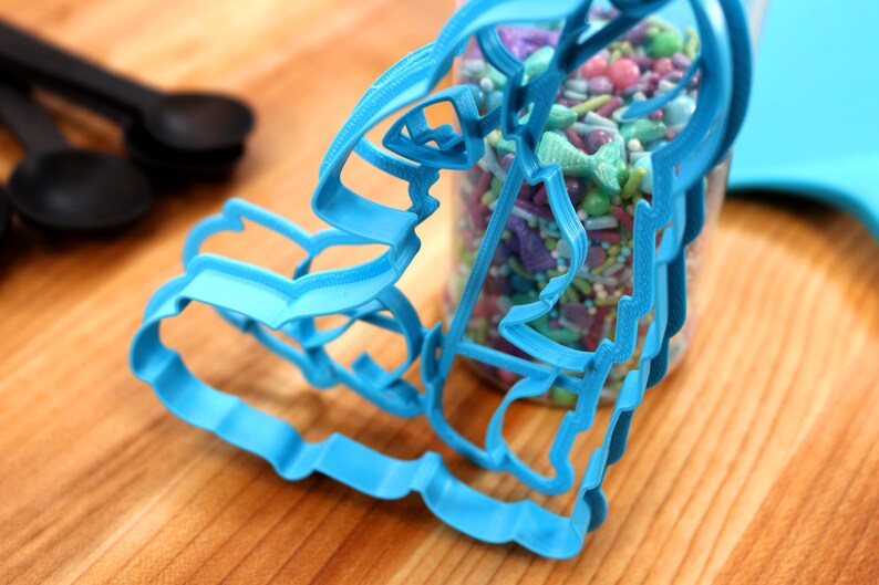 May include: A blue plastic cookie cutter shaped like a mermaid, with a jar of colourful sprinkles in the background.
