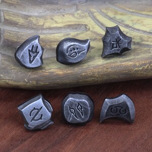 Puede incluir: Seis amuletos de metal plateado con diferentes dise&ntilde;os. Los amuletos est&aacute;n sobre una superficie de madera. Los dise&ntilde;os incluyen una mano, una huella de pata, un s&iacute;mbolo, una letra Z, dos l&iacute;neas y un c&iacute;rculo con dos puntos.