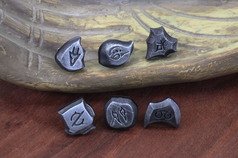 Puede incluir: Seis amuletos de metal plateado con diferentes dise&ntilde;os. Los amuletos est&aacute;n sobre una superficie de madera. Los dise&ntilde;os incluyen una mano, una huella de pata, un s&iacute;mbolo, una letra Z, dos l&iacute;neas y un c&iacute;rculo con dos puntos.