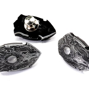 May include: Three silver and black shield-shaped pins with intricate designs. The pins have a silver finish and a black background with a silver centre. The pins are designed to be worn on clothing or bags.
