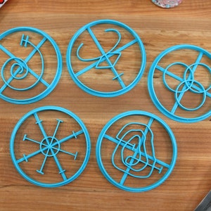 Könnte beinhalten: Fünf blaue Plastik-Ausstecher mit verschiedenen Designs. Die Designs umfassen eine Spirale, ein Kreuz mit einer geschwungenen Linie, drei überlappende Kreise, eine Sternform und eine geschwungene Linie.