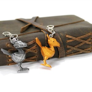 May include: Two metal keychains shaped like a bird, one silver and one gold, are attached to a brown leather journal with a braided strap.