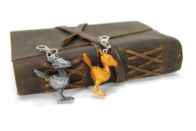 May include: Two metal keychains shaped like a bird, one silver and one gold, are attached to a brown leather journal with a braided strap.