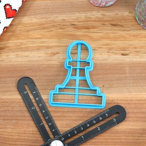 May include: Blue plastic cookie cutter shaped like a chess pawn. The cutter is on a wooden surface with a ruler and a container of sprinkles.