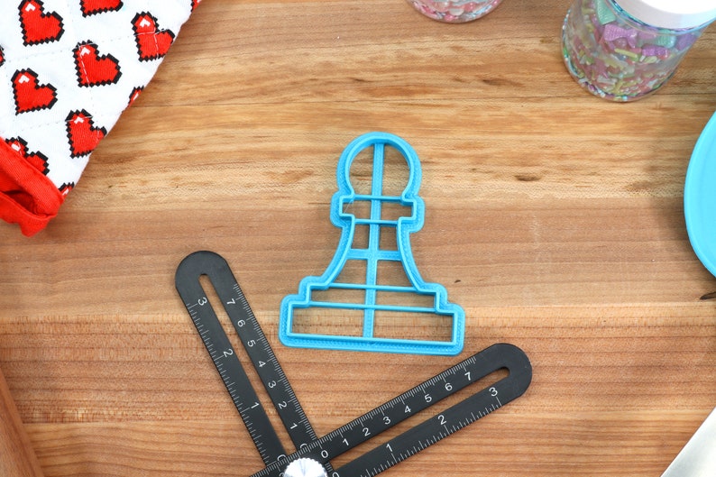 May include: Blue plastic cookie cutter shaped like a chess pawn. The cutter is on a wooden surface with a ruler and a container of sprinkles.