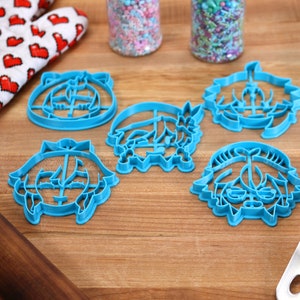 Pu&ograve; includere: Cinque tagliabiscotti in plastica blu a forma di facce dei cartoni animati. I tagliabiscotti sono su una superficie di legno con un matterello, un guanto da forno rosso e bianco e una spatola blu.