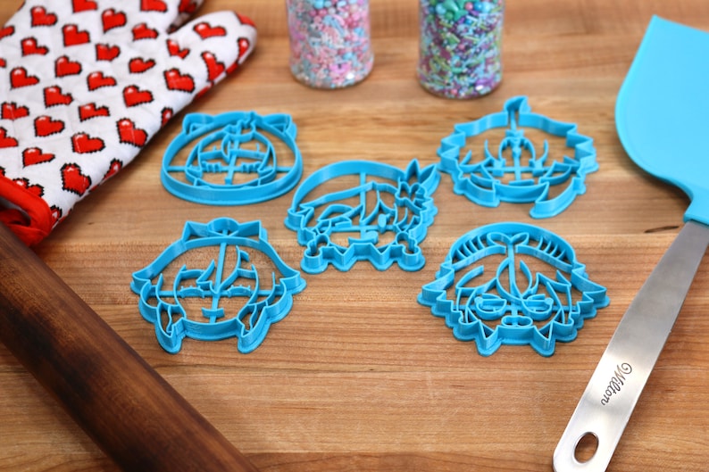 Pu&ograve; includere: Cinque tagliabiscotti in plastica blu a forma di facce dei cartoni animati. I tagliabiscotti sono su una superficie di legno con un matterello, un guanto da forno rosso e bianco e una spatola blu.