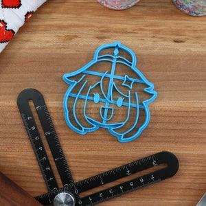 May include: A blue plastic cookie cutter shaped like a cartoon character with a star on their head. The character has a happy expression and is wearing a hat.