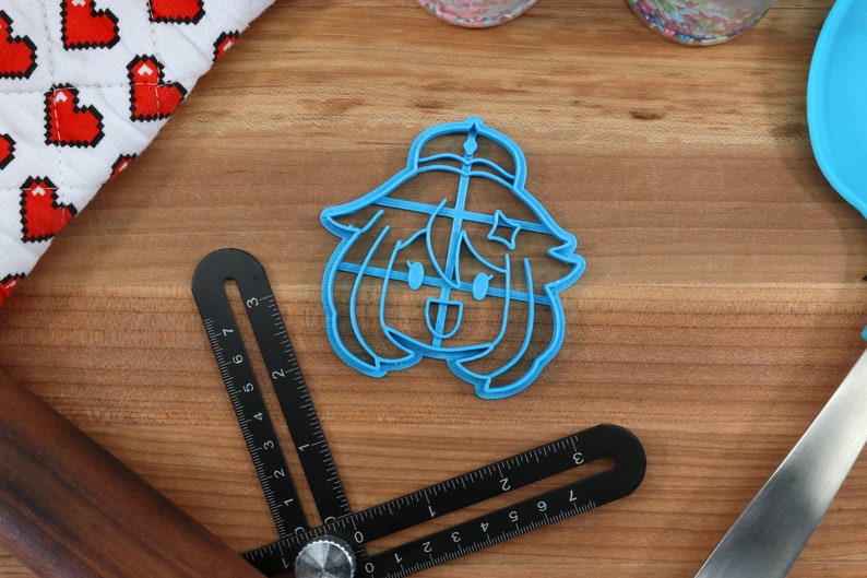 May include: A blue plastic cookie cutter shaped like a cartoon character with a star on their head. The character has a happy expression and is wearing a hat.