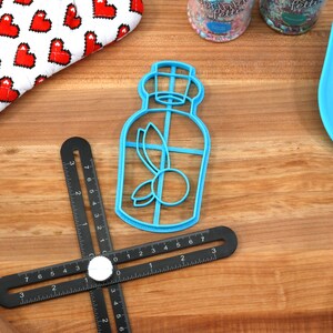 May include: A blue plastic cookie cutter shaped like a bottle with a leaf and a berry inside. The cutter is on a wooden surface with a black ruler and a blue spatula.
