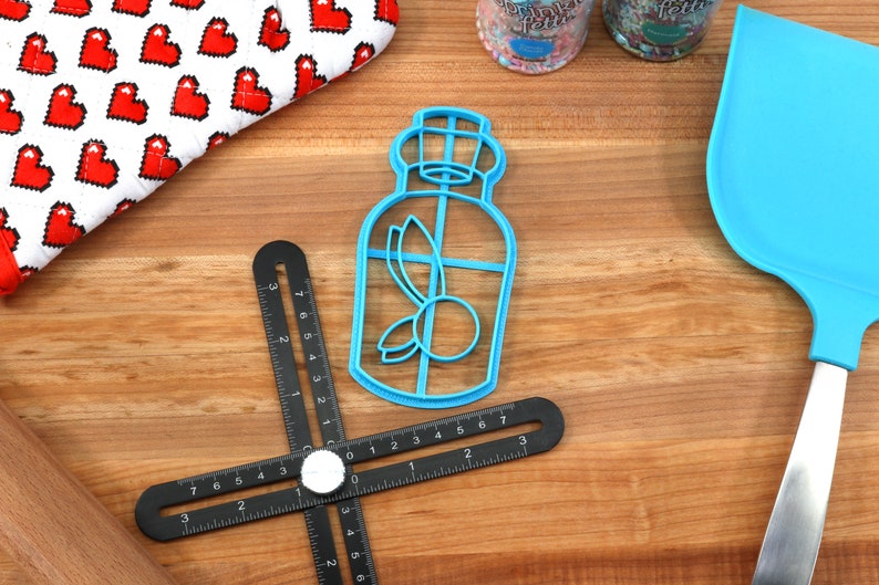 May include: A blue plastic cookie cutter shaped like a bottle with a leaf and a berry inside. The cutter is on a wooden surface with a black ruler and a blue spatula.