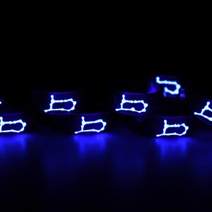 以下が含まれることがあります： 黒いシリコン製のリング8個のセット。各リングには白い光る「B」の文字がデザインされています。リングは黒い表面に一列に並べてあります。