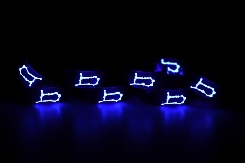 以下が含まれることがあります： 黒いシリコン製のリング8個のセット。各リングには白い光る「B」の文字がデザインされています。リングは黒い表面に一列に並べてあります。
