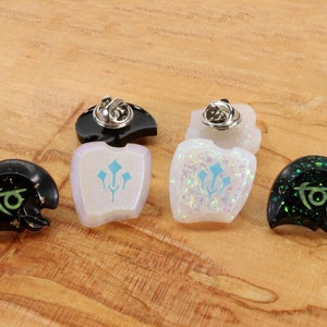 May include: Four enamel pins with different designs. Two pins are black with gold glitter and green designs. One pin is white with blue and green designs. One pin is white with iridescent glitter and blue and green designs.
