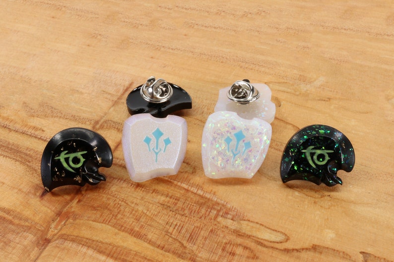 May include: Four enamel pins with different designs. Two pins are black with gold glitter and green designs. One pin is white with blue and green designs. One pin is white with iridescent glitter and blue and green designs.