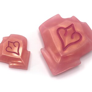 May include: Two pink, geometric-shaped objects with a heart design. The objects have a metallic sheen and are set against a white background. The heart design is outlined in a darker pink color. The objects appear to be decorative or functional.