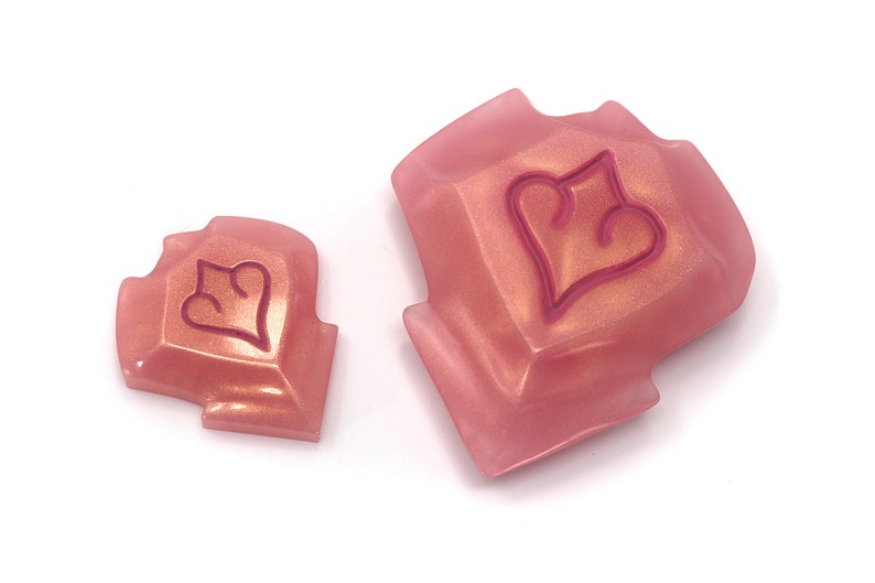 May include: Two pink, geometric-shaped objects with a heart design. The objects have a metallic sheen and are set against a white background. The heart design is outlined in a darker pink color. The objects appear to be decorative or functional.