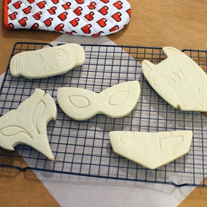 May include: Unbaked cookies in various shapes, including a mask, fox, and owl, rest on a black wire cooling rack. A red and white heart-patterned oven mitt is in the background, suggesting a baking theme.
