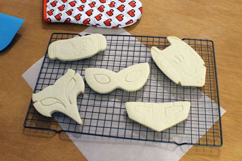 May include: Unbaked cookies in various shapes, including a mask, fox, and owl, rest on a black wire cooling rack. A red and white heart-patterned oven mitt is in the background, suggesting a baking theme.