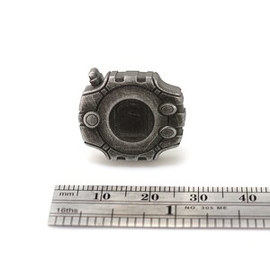 May include: A silver-colored metal pin with a circular design featuring a raised, textured border and a central circular area with a dark gray background. The pin is approximately 2 cm in diameter.