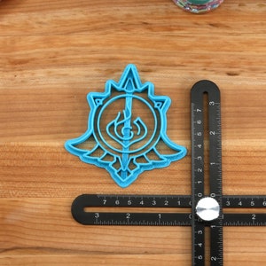 May include: A blue plastic cookie cutter shaped like a stylized sword and shield with a flame design. The cutter is on a wooden surface with a ruler.