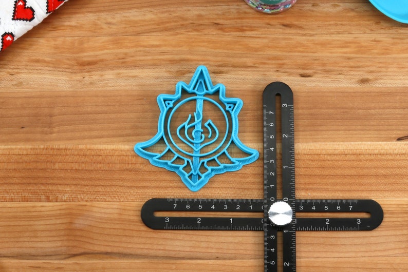 May include: A blue plastic cookie cutter shaped like a stylized sword and shield with a flame design. The cutter is on a wooden surface with a ruler.