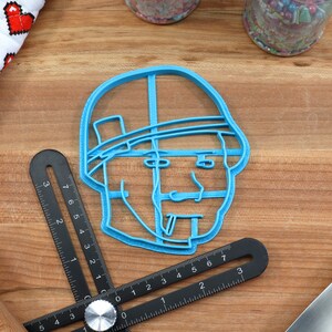 May include: A blue plastic cookie cutter shaped like a person's head wearing a hat. The cookie cutter is on a wooden surface.