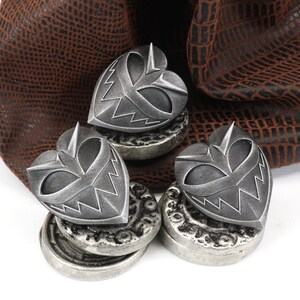 May include: Three silver heart-shaped charms with a menacing face design, resting on top of stacks of silver coins. The charms are sitting on a brown leather pouch.