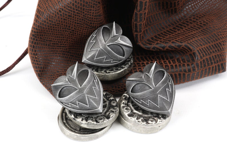 May include: Three silver heart-shaped charms with a menacing face design, resting on top of stacks of silver coins. The charms are sitting on a brown leather pouch.