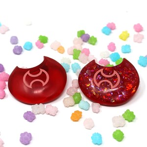 May include: Two red crescent moon shaped resin charms with a pink symbol in the center. The charms are surrounded by colorful candy sprinkles.