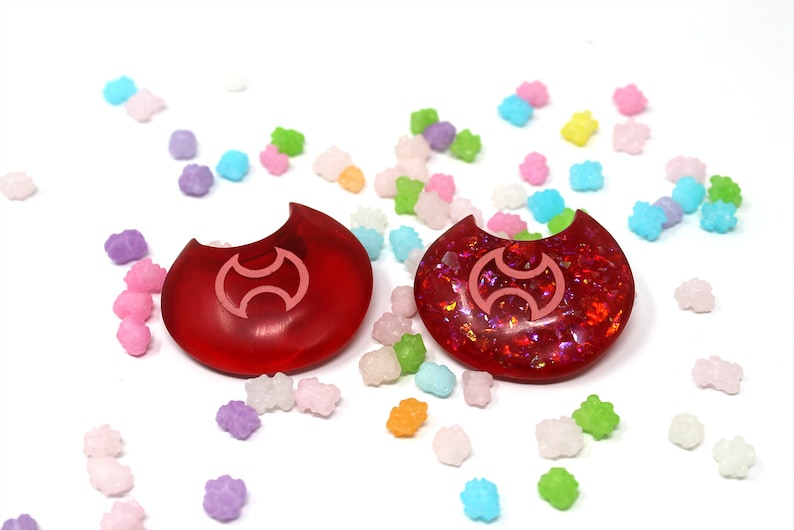 May include: Two red crescent moon shaped resin charms with a pink symbol in the center. The charms are surrounded by colorful candy sprinkles.