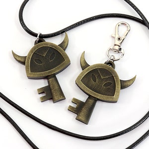 May include: Two gold-toned key pendants with a horned helmet design. One pendant is attached to a black cord necklace, the other to a black cord keychain.