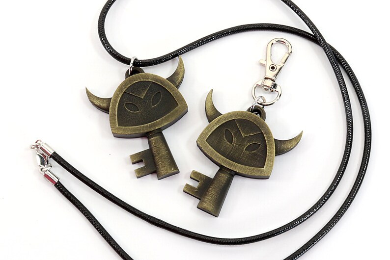 May include: Two gold-toned key pendants with a horned helmet design. One pendant is attached to a black cord necklace, the other to a black cord keychain.