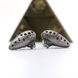 Pu&ograve; includere: Due orecchini color argento a forma di ocarina, con fori multipli. Gli orecchini hanno un aspetto strutturato e invecchiato. Una forma geometrica sfocata &egrave; sullo sfondo. Gli orecchini sono su una superficie bianca.
