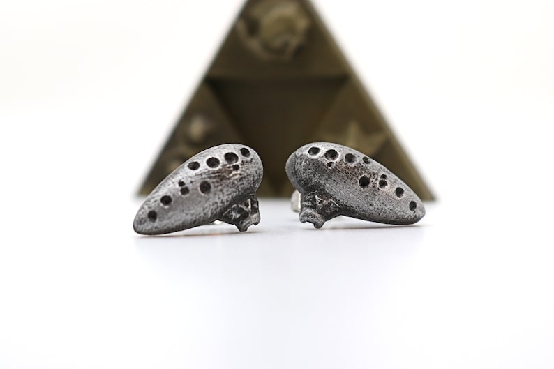 Pu&ograve; includere: Due orecchini color argento a forma di ocarina, con fori multipli. Gli orecchini hanno un aspetto strutturato e invecchiato. Una forma geometrica sfocata &egrave; sullo sfondo. Gli orecchini sono su una superficie bianca.