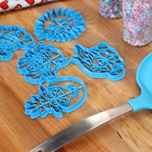 May include: A set of five blue plastic cookie cutters shaped like different cartoon characters on a wooden surface. The cutters are arranged in a semi-circle with a blue spatula and two jars of sprinkles in the background.