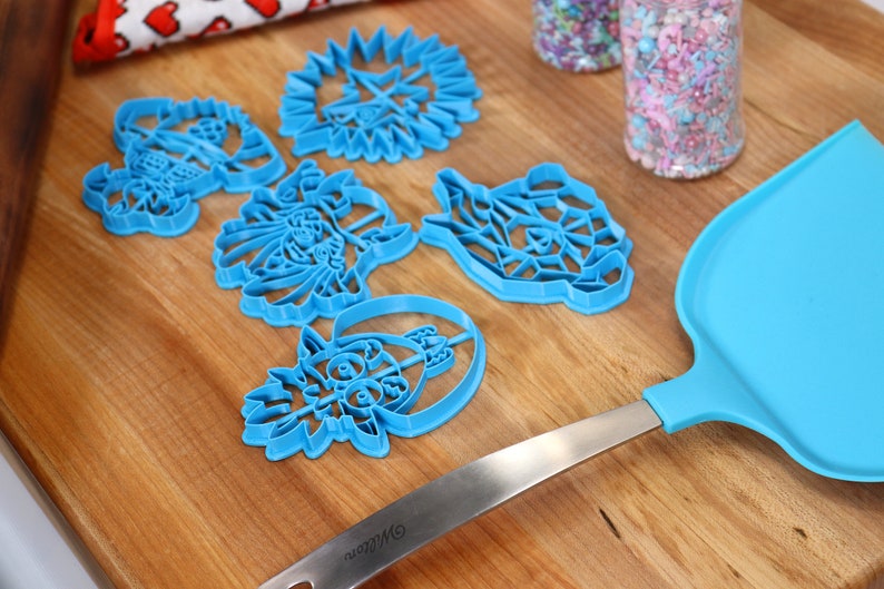 May include: A set of five blue plastic cookie cutters shaped like different cartoon characters on a wooden surface. The cutters are arranged in a semi-circle with a blue spatula and two jars of sprinkles in the background.