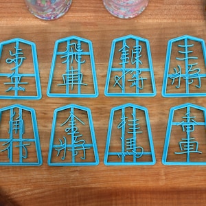 May include: Eight light blue cookie cutters shaped like Japanese Shogi pieces, each with a different character. The cutters are on a wooden surface with a blue spatula and rolling pin visible.