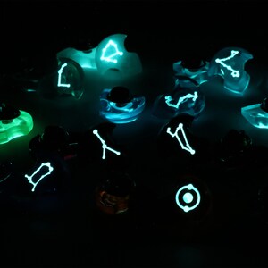 May include: A collection of small, clear glass ornaments with glowing constellations inside. Each ornament features a different constellation, such as Gemini, Cancer, and Leo. The ornaments are arranged on a dark surface, creating a celestial and magical atmosphere.