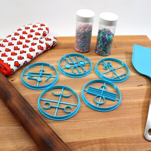 May include: A wooden rolling pin, a blue oven mitt with red hearts, five blue cookie cutters, two jars of sprinkles, and a blue spatula on a wooden cutting board.
