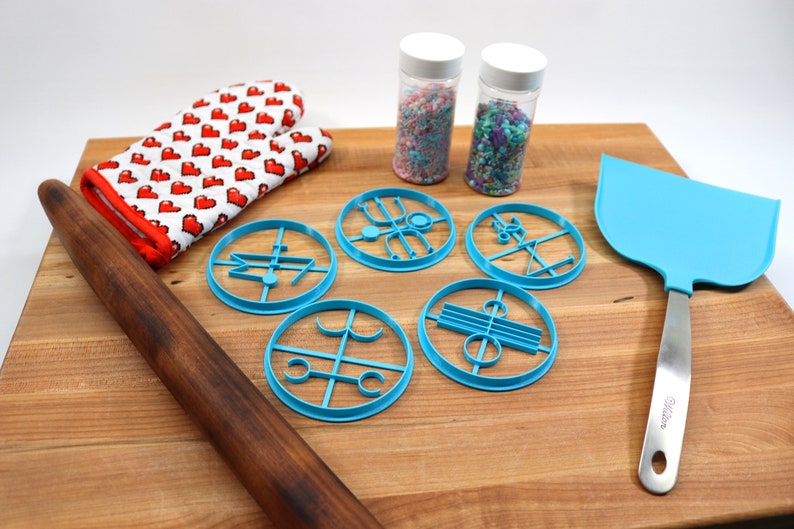 May include: A wooden rolling pin, a blue oven mitt with red hearts, five blue cookie cutters, two jars of sprinkles, and a blue spatula on a wooden cutting board.