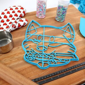 May include: A blue plastic cookie cutter shaped like a fox's face on a wooden surface with a rolling pin, measuring tape, a spatula, a measuring cup, and two jars of sprinkles.
