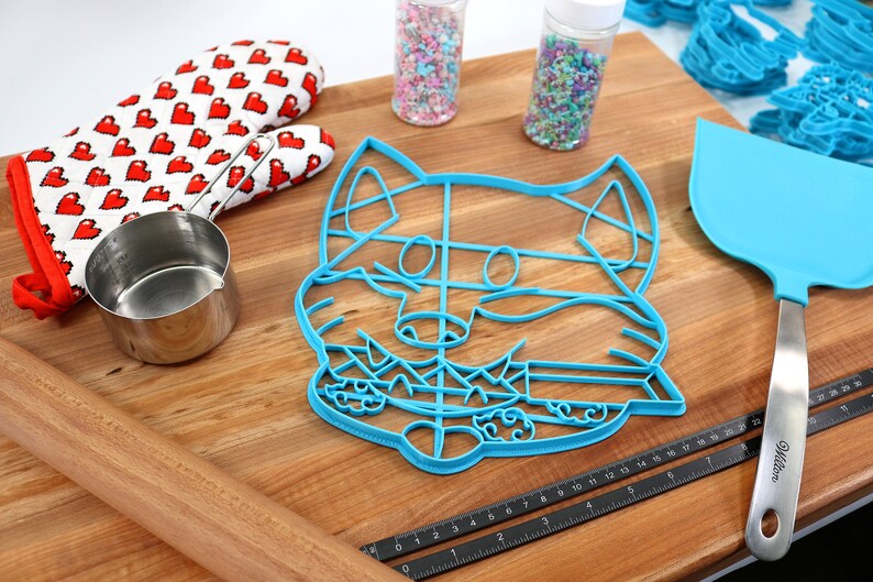 May include: A blue plastic cookie cutter shaped like a fox's face on a wooden surface with a rolling pin, measuring tape, a spatula, a measuring cup, and two jars of sprinkles.